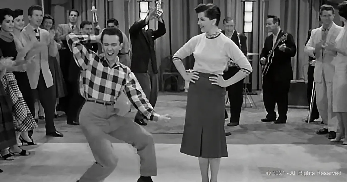 A man in a checkered shirt dances enthusiastically while a woman in a skirt watches, surrounded by a lively crowd.