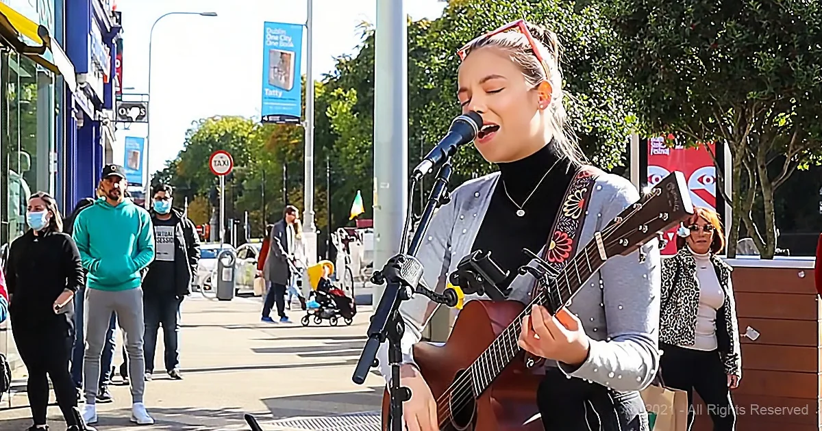 A woman sings passionately while playing guitar on a city street, surrounded by pedestrians and urban scenery.