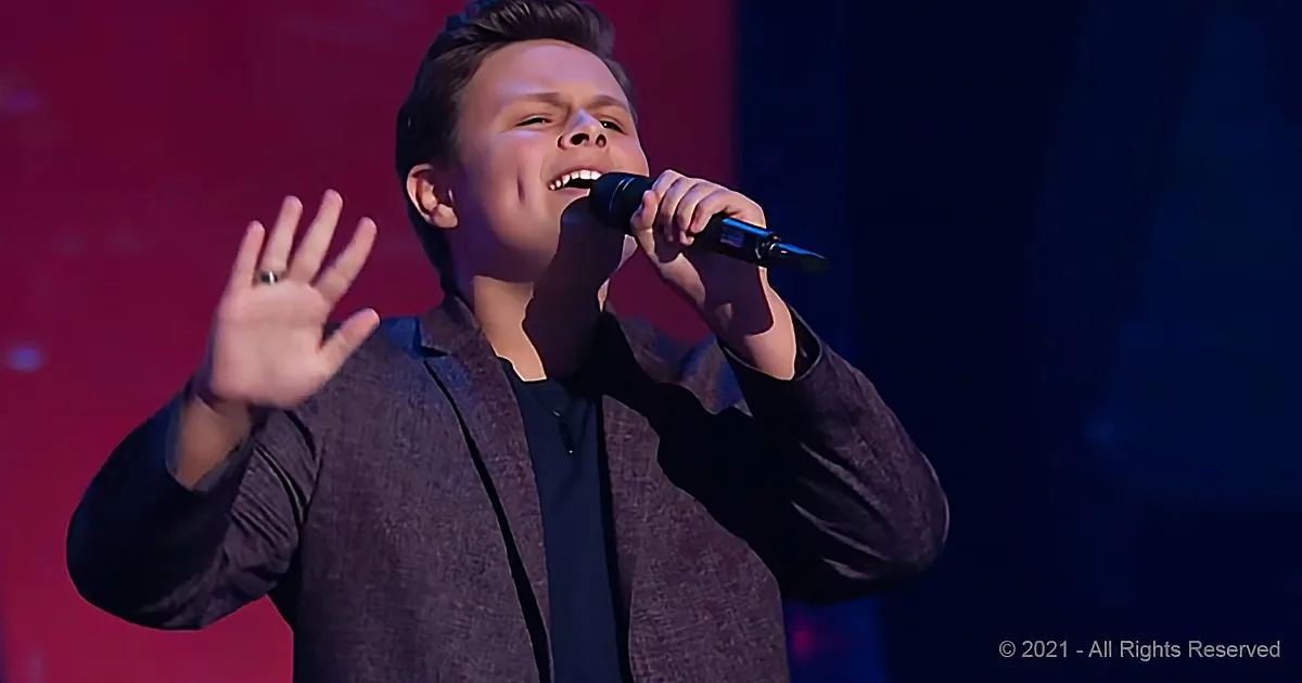 A young person in a suit sings into a microphone, gesturing with one hand raised.