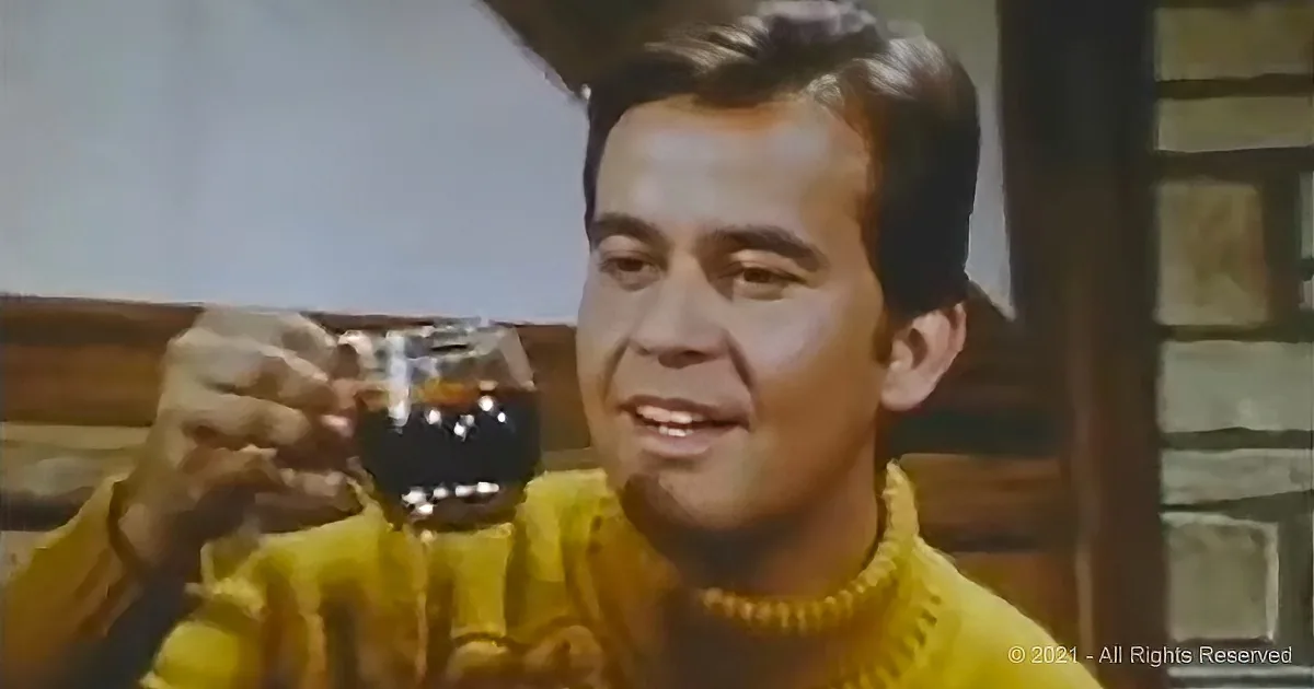 A man in a yellow sweater holds a glass of dark liquid, smiling.