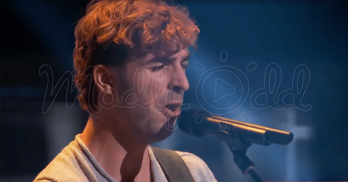 A person with curly hair, wearing a white shirt, is singing into a microphone on stage.