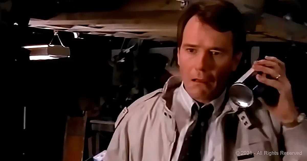 A man in a trench coat holding a flashlight, looking concerned, in a dimly lit room.