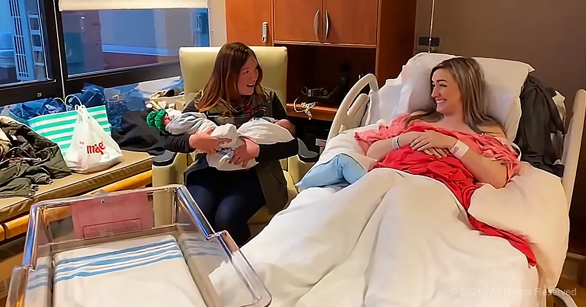 A woman in a hospital bed holds a newborn baby while another woman sits beside her, both smiling.