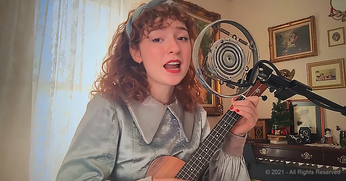 A woman sings into a microphone while playing a ukulele in a cozy room with framed pictures.