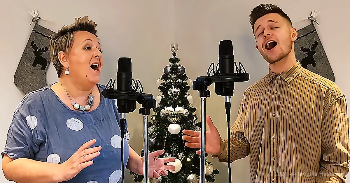 A woman and man sing into microphones in front of a decorated Christmas tree.