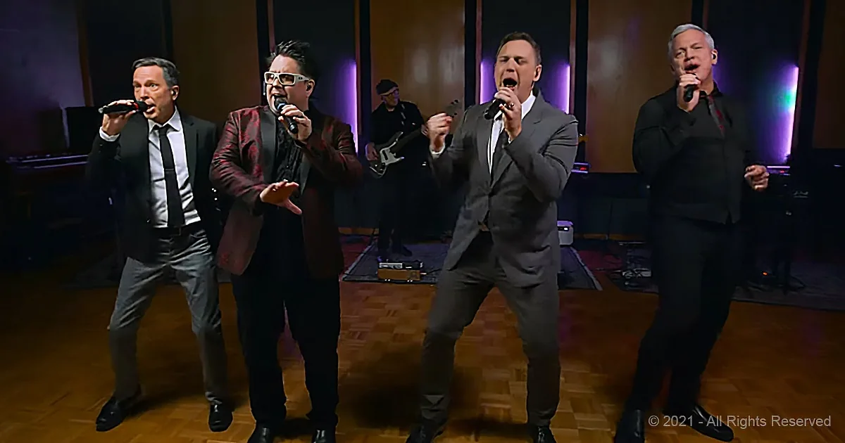 Four men in suits sing passionately on stage with a guitarist in the background.