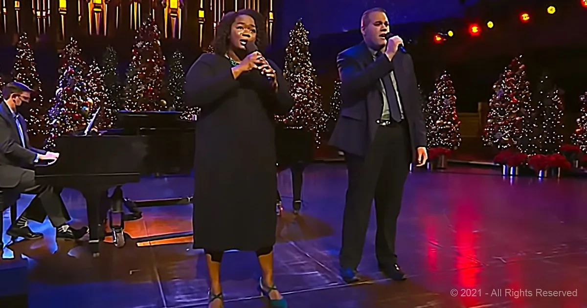A woman and man sing on stage with Christmas trees and a pianist in the background.