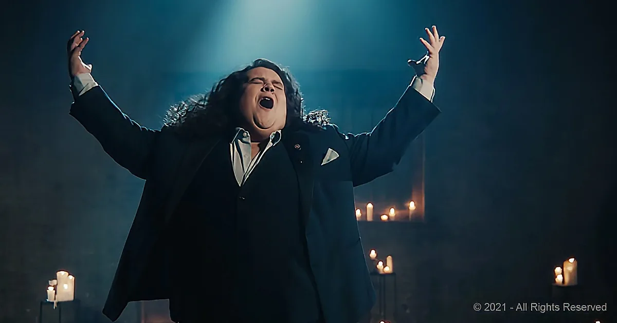 A man in a suit sings passionately with his arms raised, surrounded by candles and dramatic lighting.
