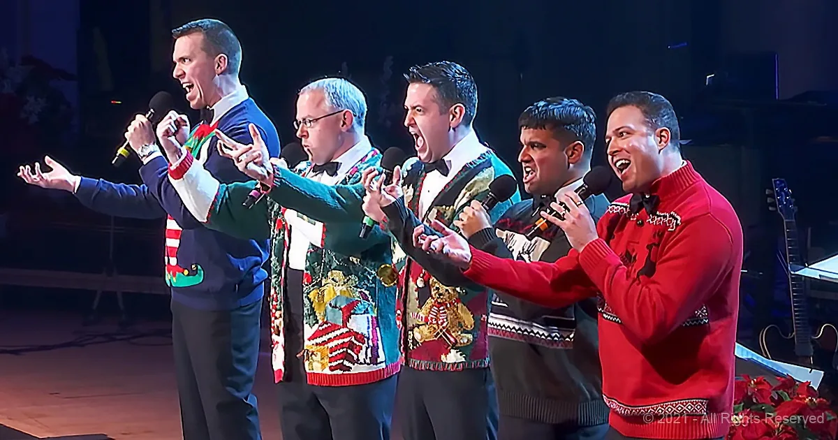 A group of men in festive sweaters sing on stage, holding microphones and expressing enthusiasm.