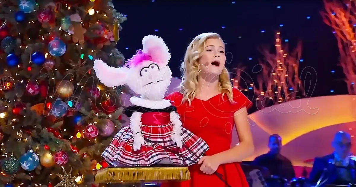 A woman in a red dress sings on stage with a puppet, a Christmas tree adorned with ornaments, and a band in the background.