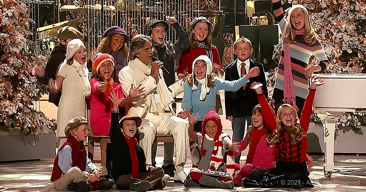 A man in white sings surrounded by children in festive winter attire, with Christmas trees and decorations in the background.