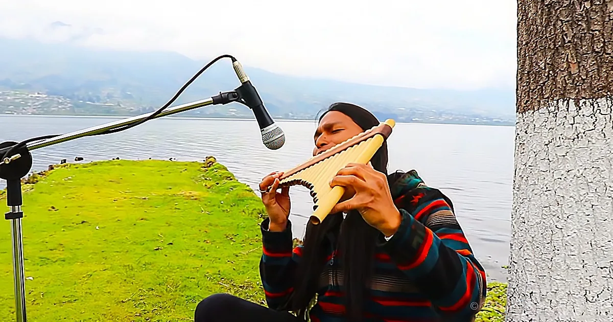 A person plays a pan flute near a body of water, with a microphone capturing the sound.