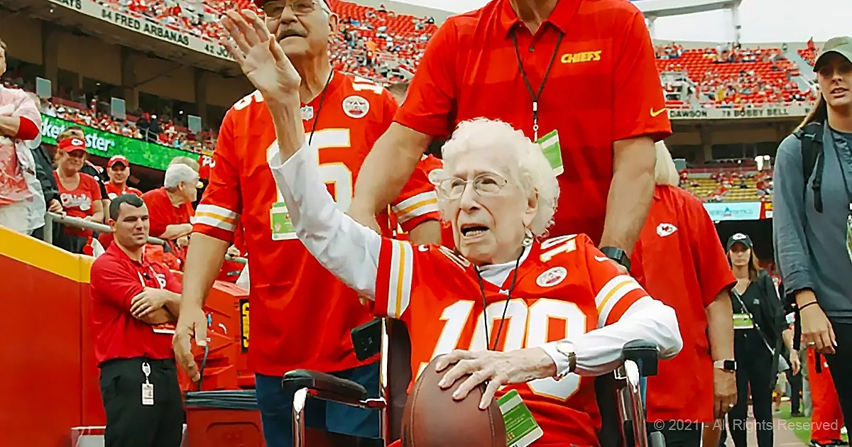 An elderly woman in a Chiefs jersey celebrates at a football game, surrounded by fans.