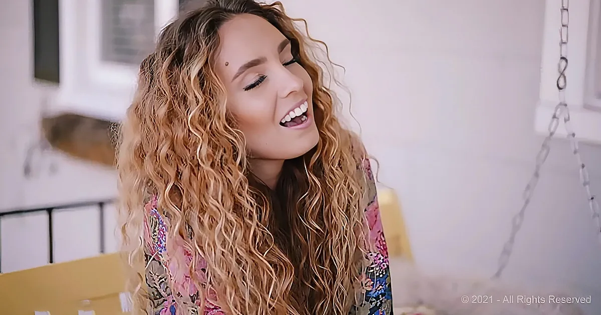 A woman with long, curly blonde hair smiles brightly, wearing a floral top, in a softly lit room.