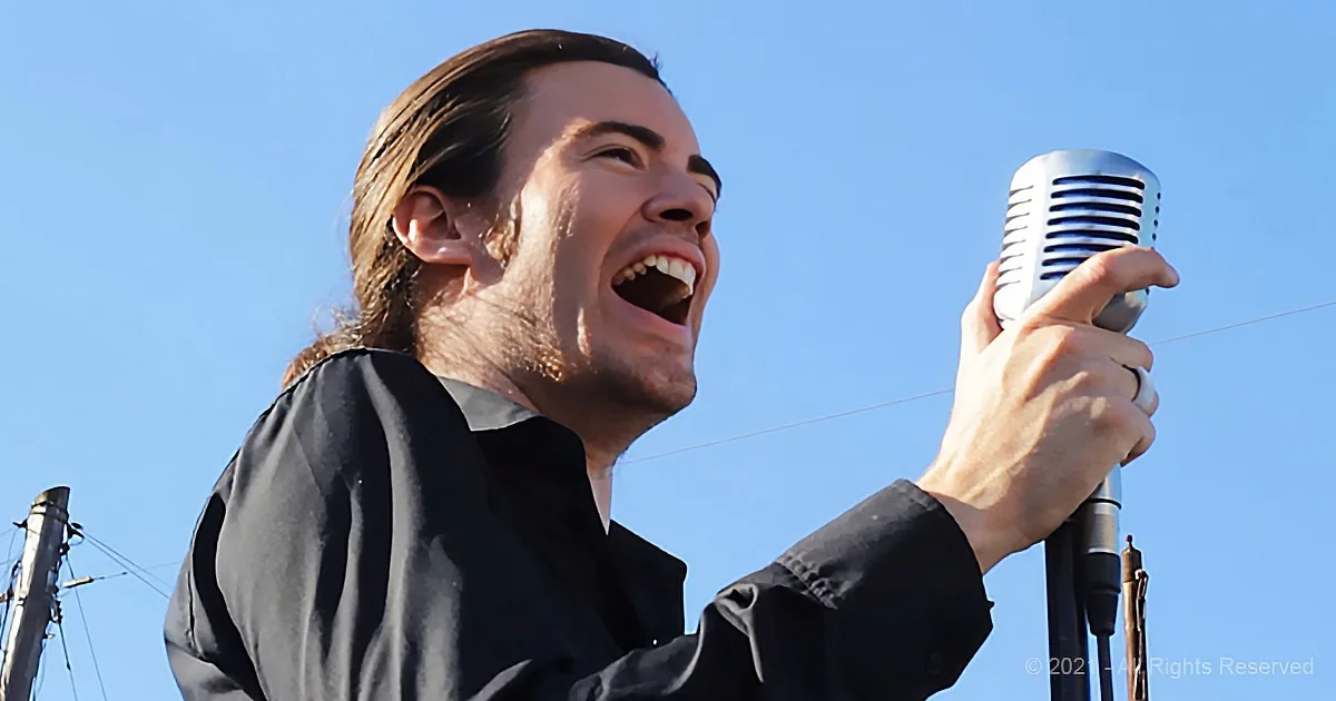 A man in a black shirt sings passionately into a vintage microphone against a clear blue sky.