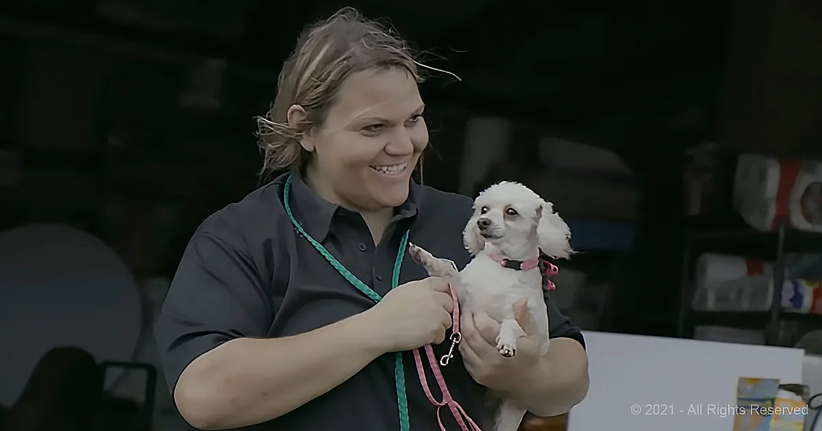 A woman holds a small white dog with a pink collar, smiling, in what appears to be a grooming or competition setting.