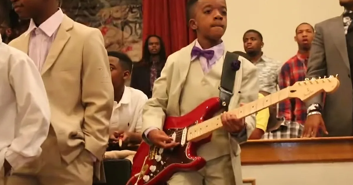 A young boy in a suit plays a red electric guitar in front of a group of people.