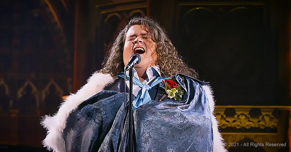 A person in a blue robe with a red rose and white fur trim sings passionately into a microphone.