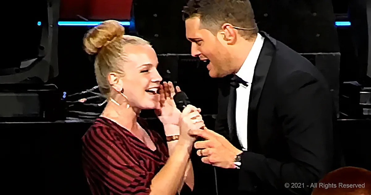 A man in a tuxedo and a woman in a striped dress share a joyful moment on stage, holding hands and smiling.