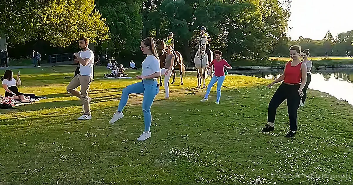 People practicing dance moves on grass near a lake with horses in the background.