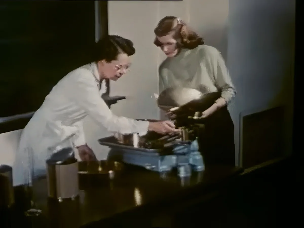 A woman in a white lab coat leans over a counter, interacting with another woman in a green blouse.