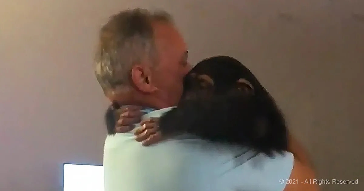 A man hugs a small black animal, possibly a baby monkey, with a light blue shirt.