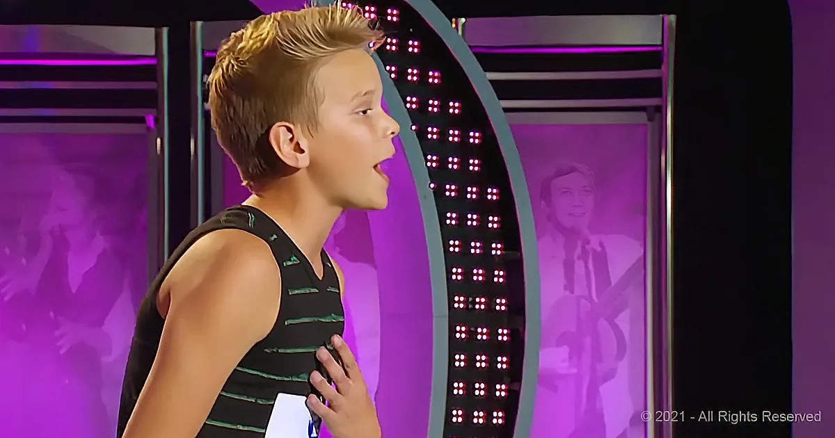 A young boy in a black tank top sings on stage with a purple-lit backdrop and a cello silhouette.
