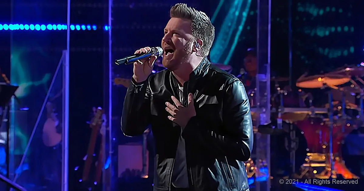 A man in a black leather jacket sings passionately into a microphone on stage.