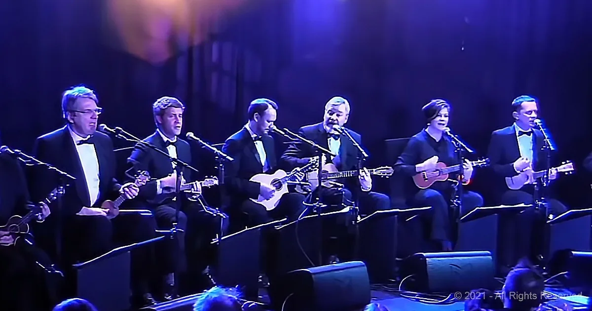 A group of musicians in tuxedos play ukuleles and sing on stage under blue lighting.