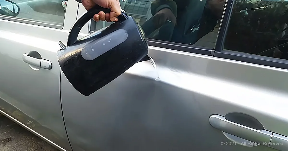 A hand holds a black kettle, pouring water onto a silver car door.