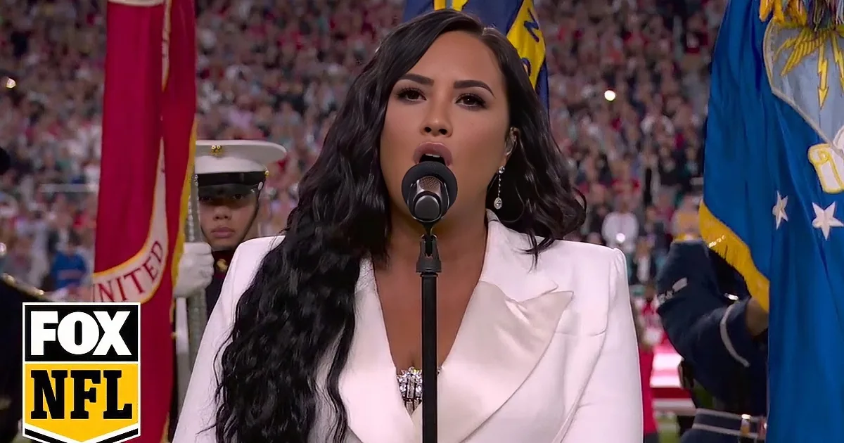 A woman in a white suit sings into a microphone, surrounded by flags and a crowd.