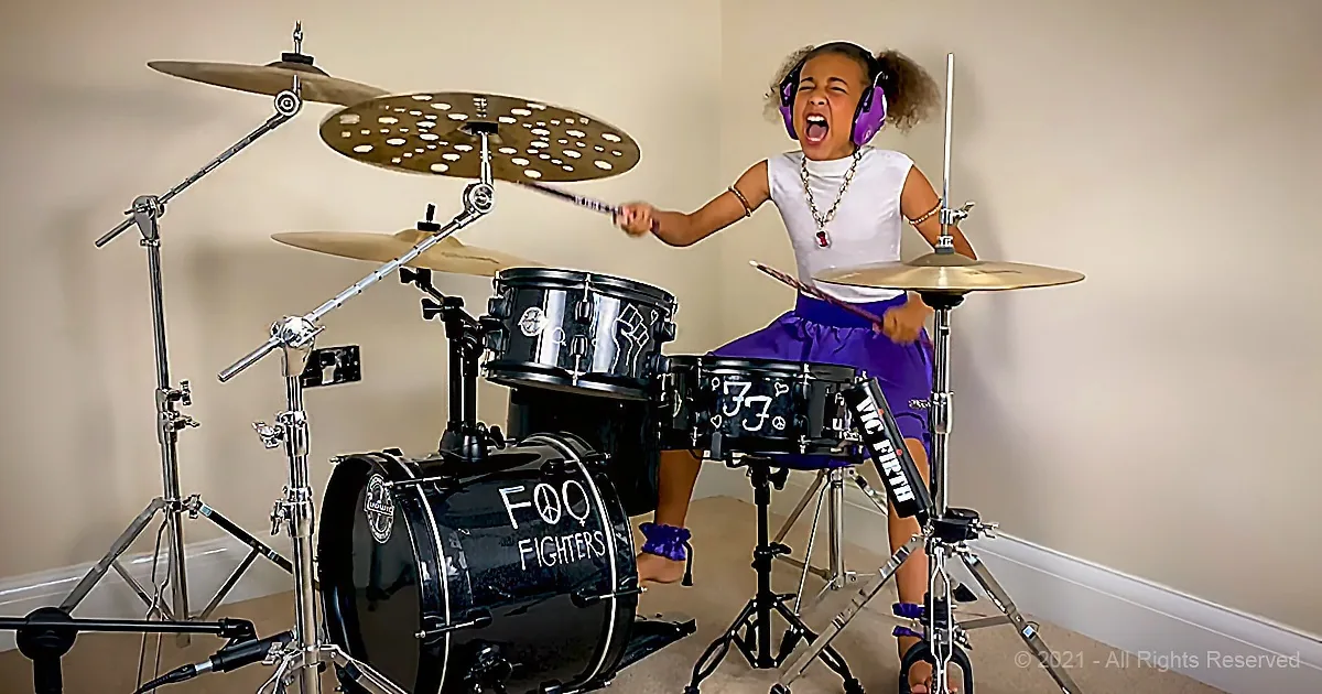 A young girl wearing purple headphones plays a Foo Fighters drum set with enthusiasm.