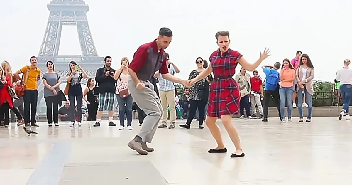 A man and woman dance energetically in front of the Eiffel Tower, surrounded by onlookers.