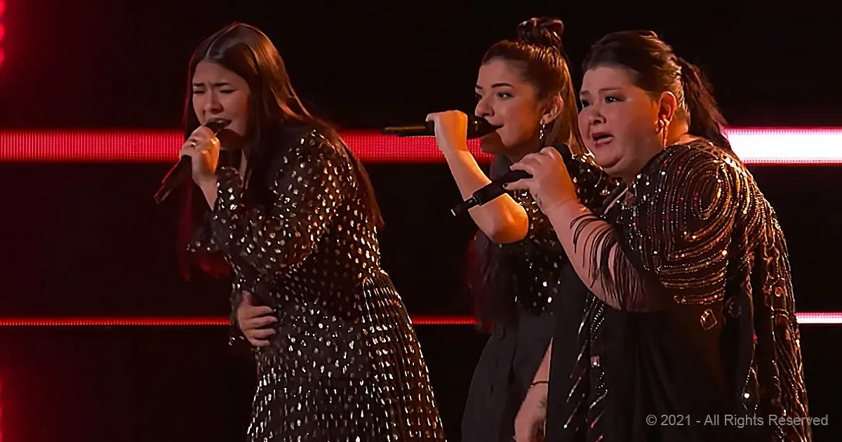 Three women in sequined dresses sing into microphones on a stage with red lights.