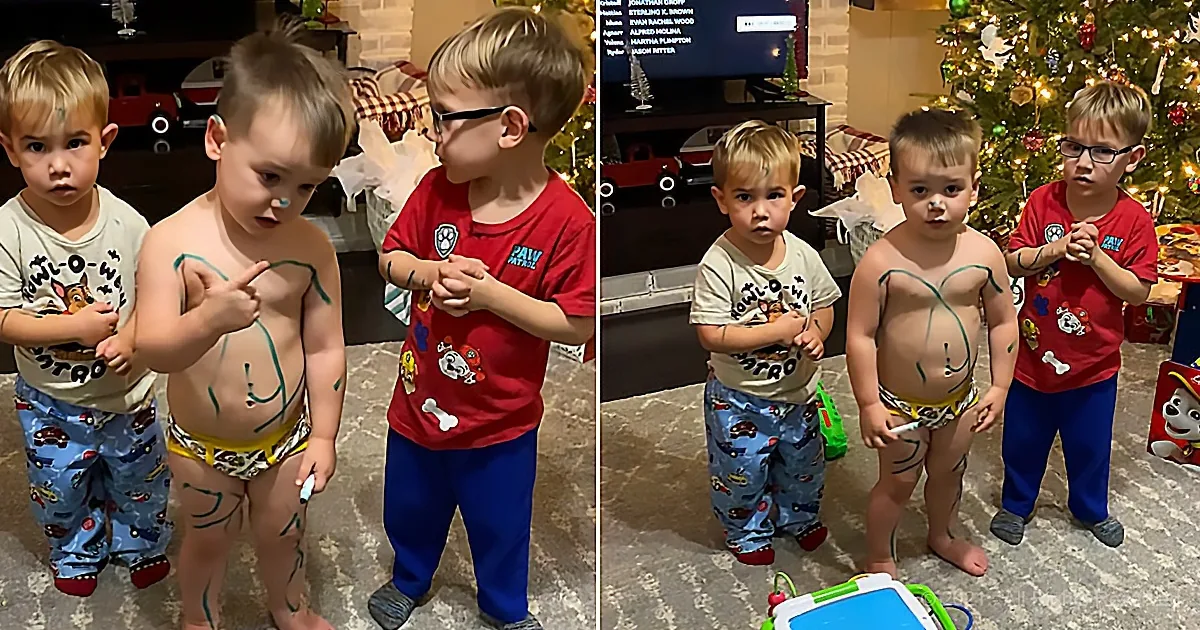 Three young boys in pajamas and underwear with green paint on their chests stand in front of a Christmas tree.