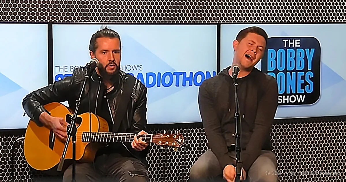 Two men, one playing guitar, the other singing, in front of "The Bobby Bones Show" backdrop.