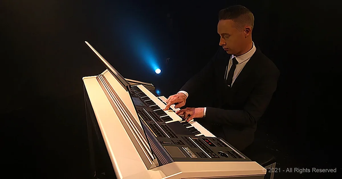 A man in a suit plays a keyboard under a spotlight in a dark setting.