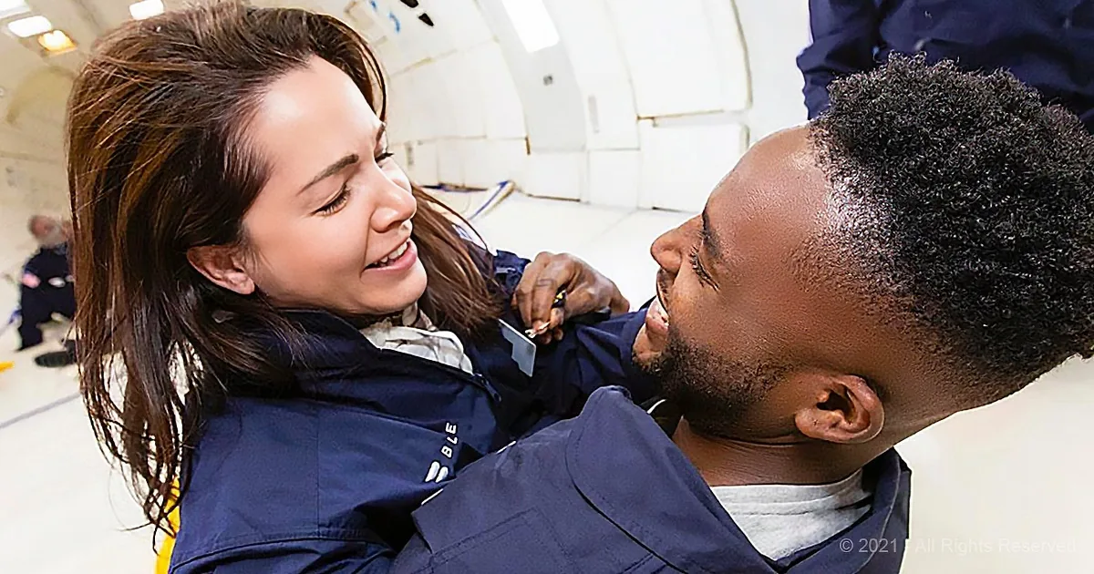 A man in a blue uniform holds a woman in a blue jacket, both smiling, in a white, industrial setting.