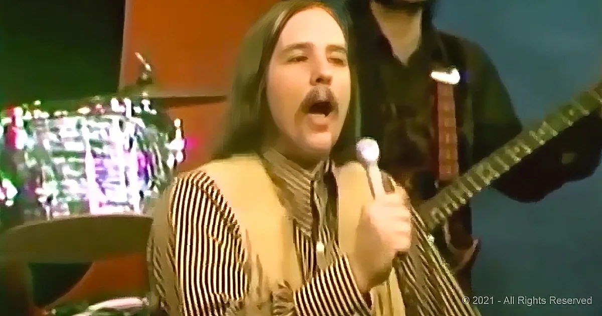 A man with long hair and a mustache sings into a microphone while playing a guitar.