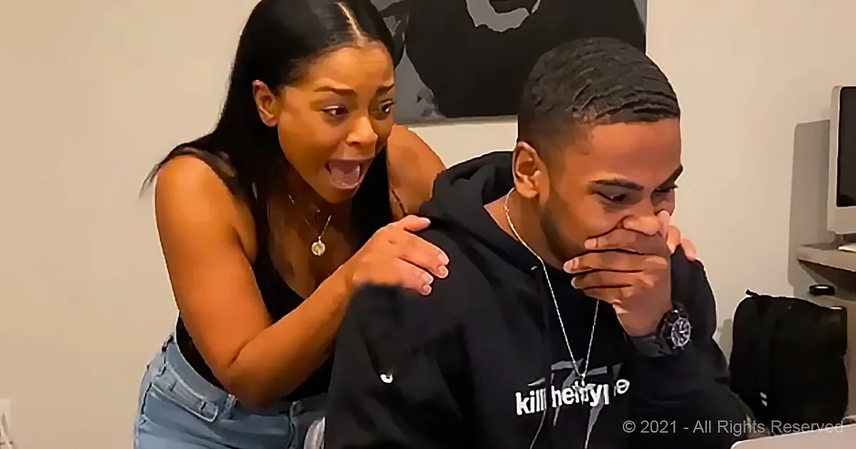 A woman leans over a man in a hoodie, both reacting to something off-screen.