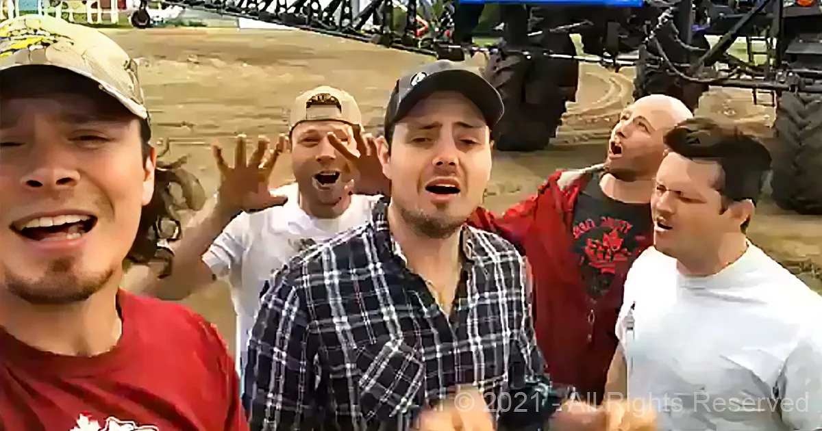 A man in a plaid shirt is surrounded by others, all appearing to shout or sing, with a sandy background and equipment...