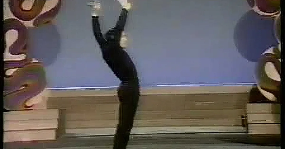A person in a black outfit balances a ball on their head while standing on a stage with a colorful background.