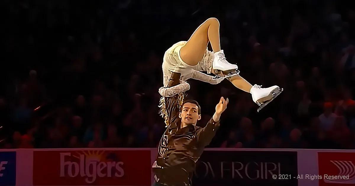 A figure skater in white is being lifted by another skater in a dramatic pose.