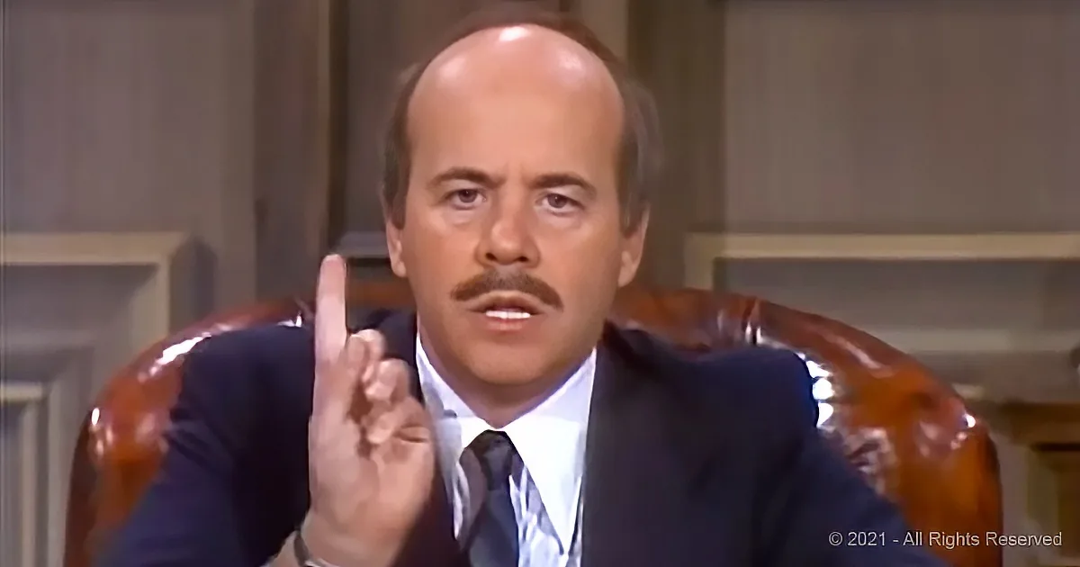 A man in a suit and tie sits in a leather chair, gesturing with his hand.