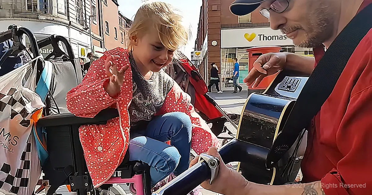 A young girl in a red coat smiles as a man plays guitar, both seated on a stroller in a sunny urban setting.