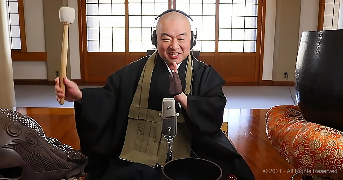 A man in traditional attire holds a drumstick, sits in front of a microphone, and gestures with his hands.