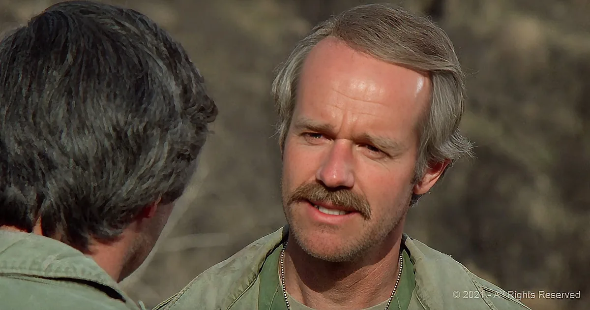 A man with gray hair and a mustache, wearing a green shirt, is speaking to another man with dark hair.