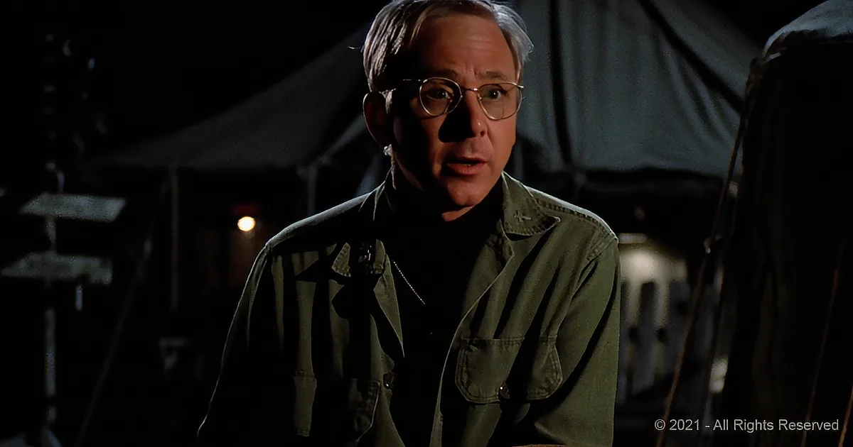 A man in a green military-style jacket stands in a dimly lit setting, possibly a camp or military base.