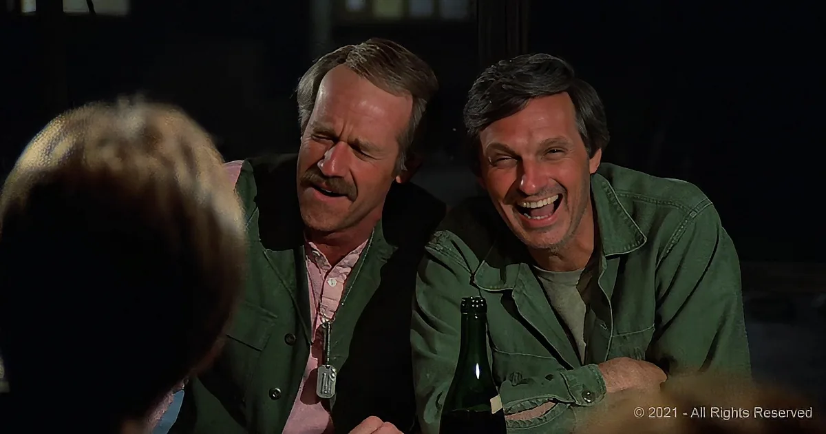 Two men in green jackets are laughing at a table, one holding a beer bottle.