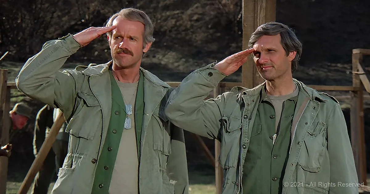 Two men in military uniforms salute, standing outdoors with a third person in the background.